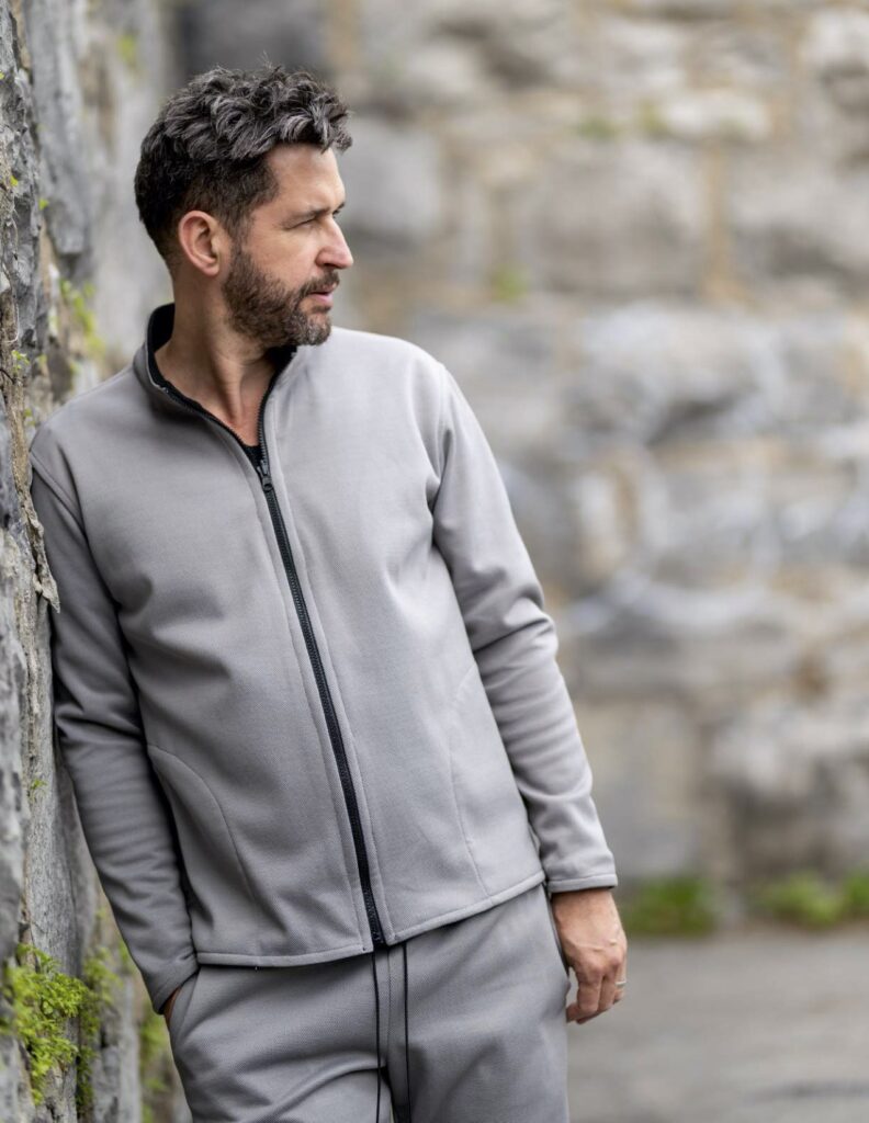 Male model wears a gray tracksuite while leaning against a stone wall
