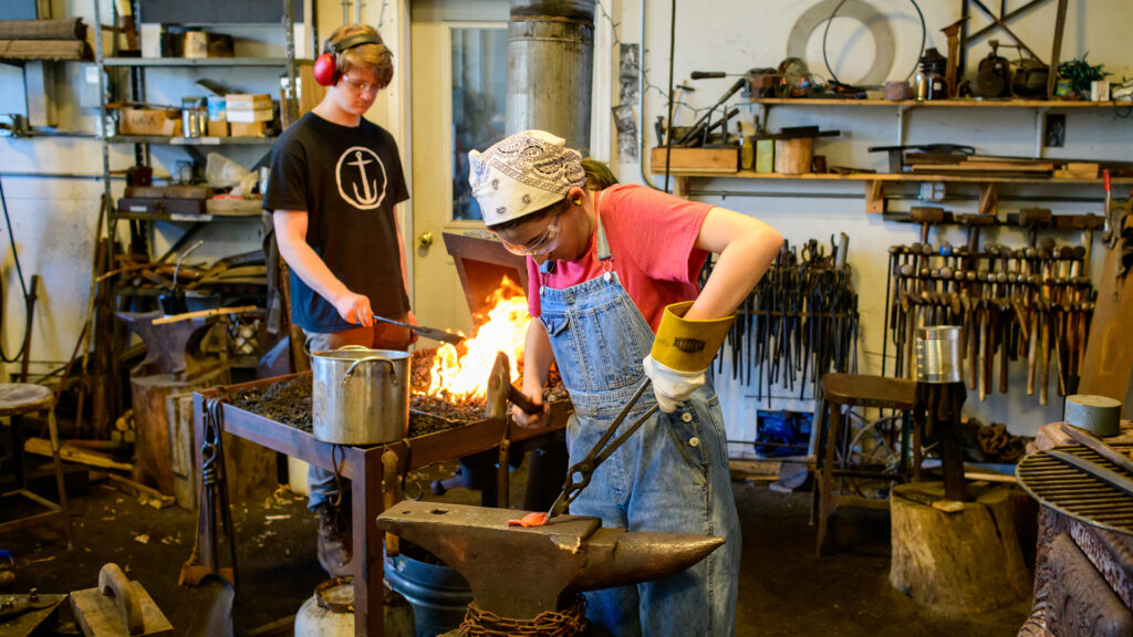 Image of teen boy and girl working in a blacksmithing shop.